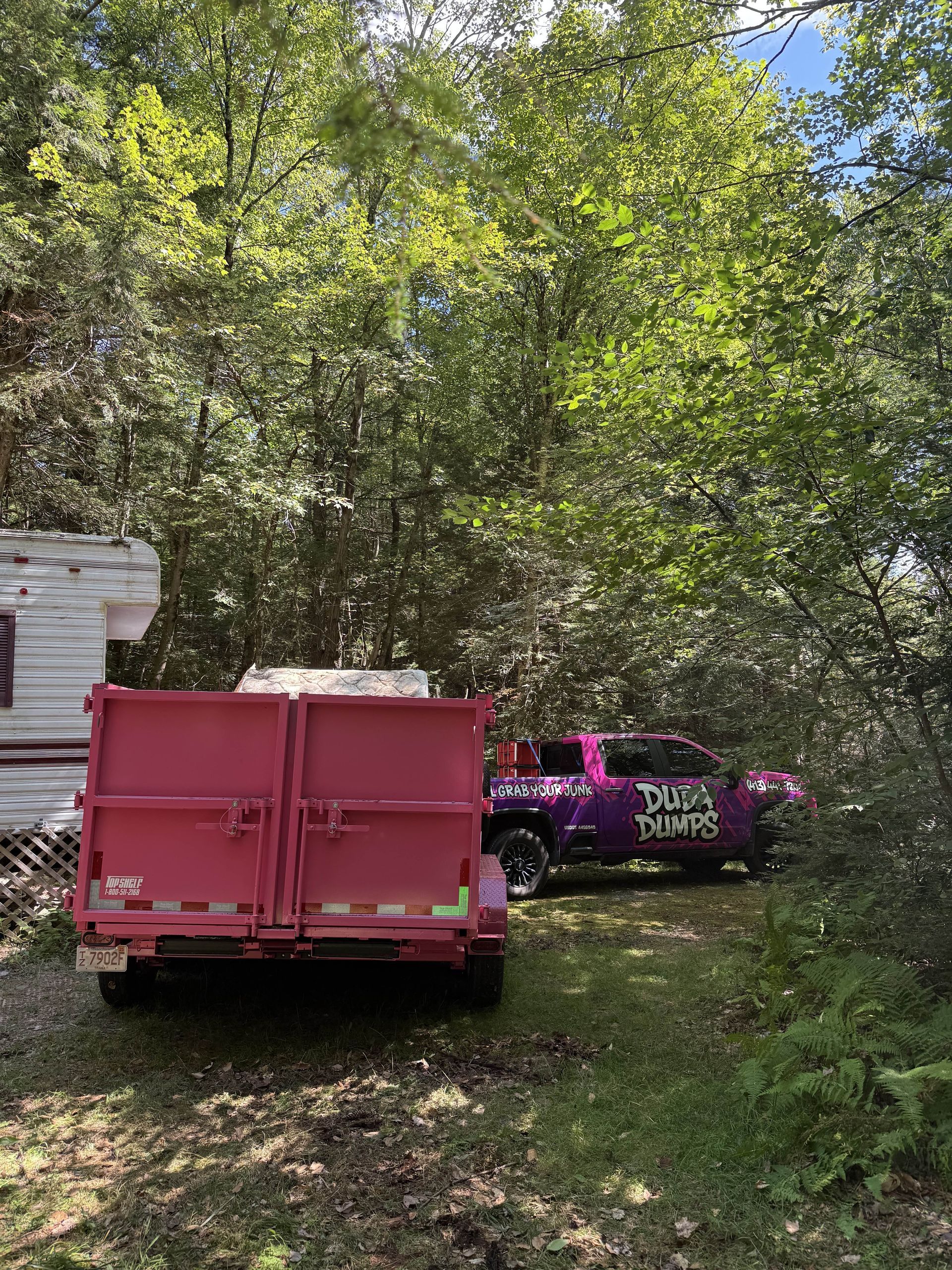 A white truck is towing a dump trailer with a tree in the background.