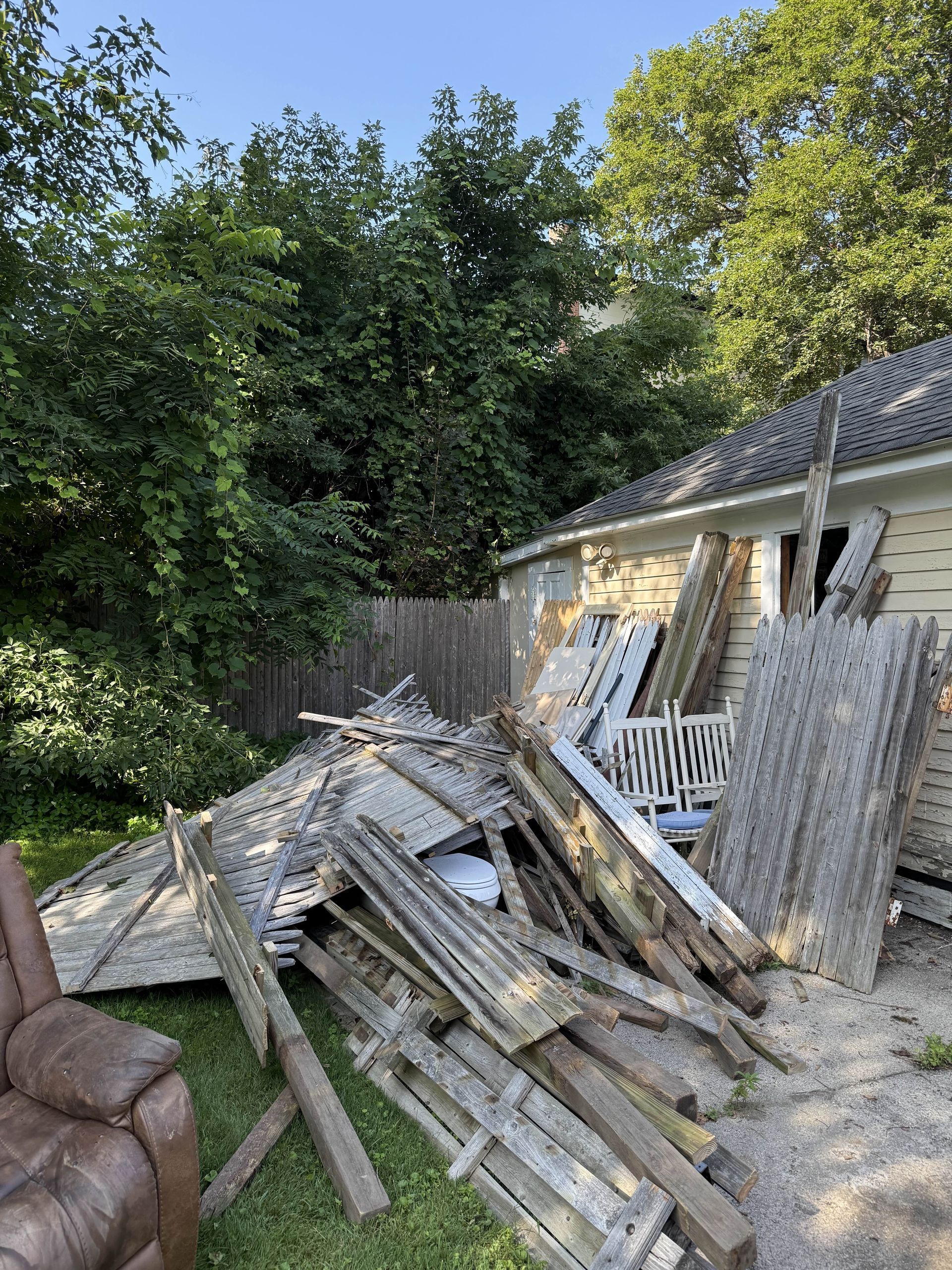 Pile of broken wooden debris near a house with trees in the background.