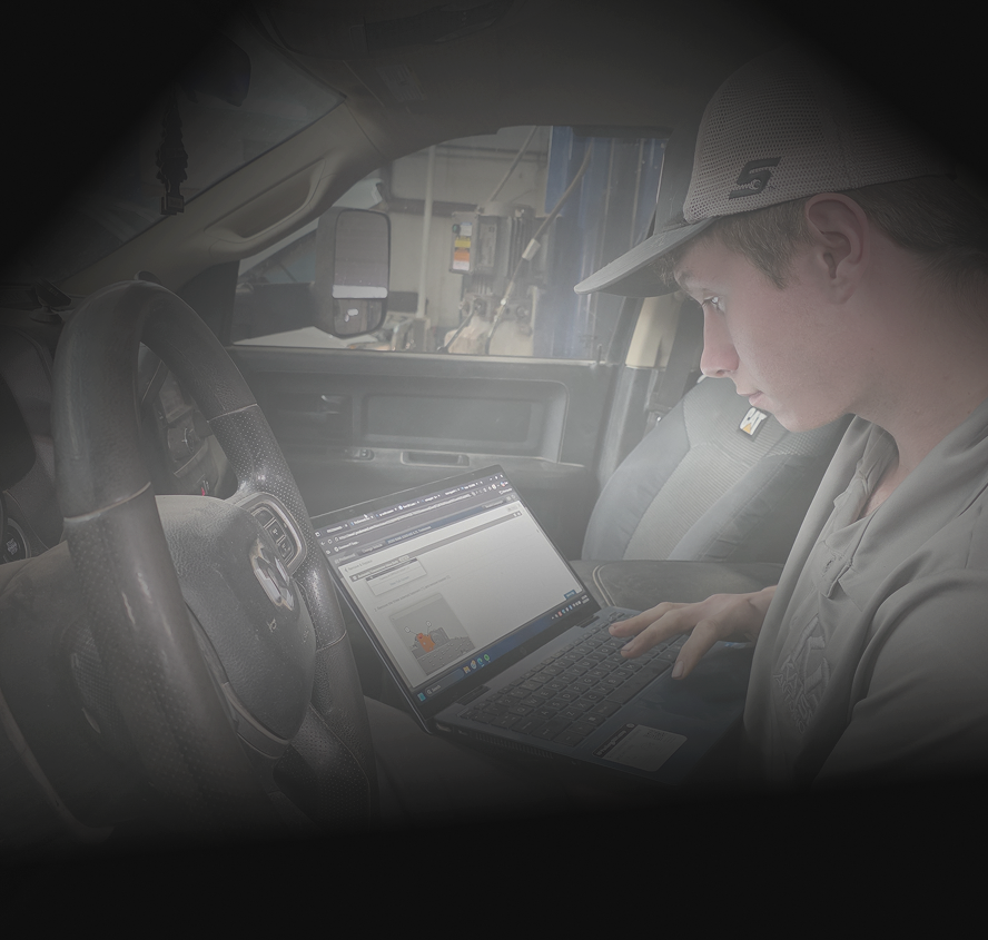 Mechanic in a garage working on a car engine, wearing a cap and holding a tool.