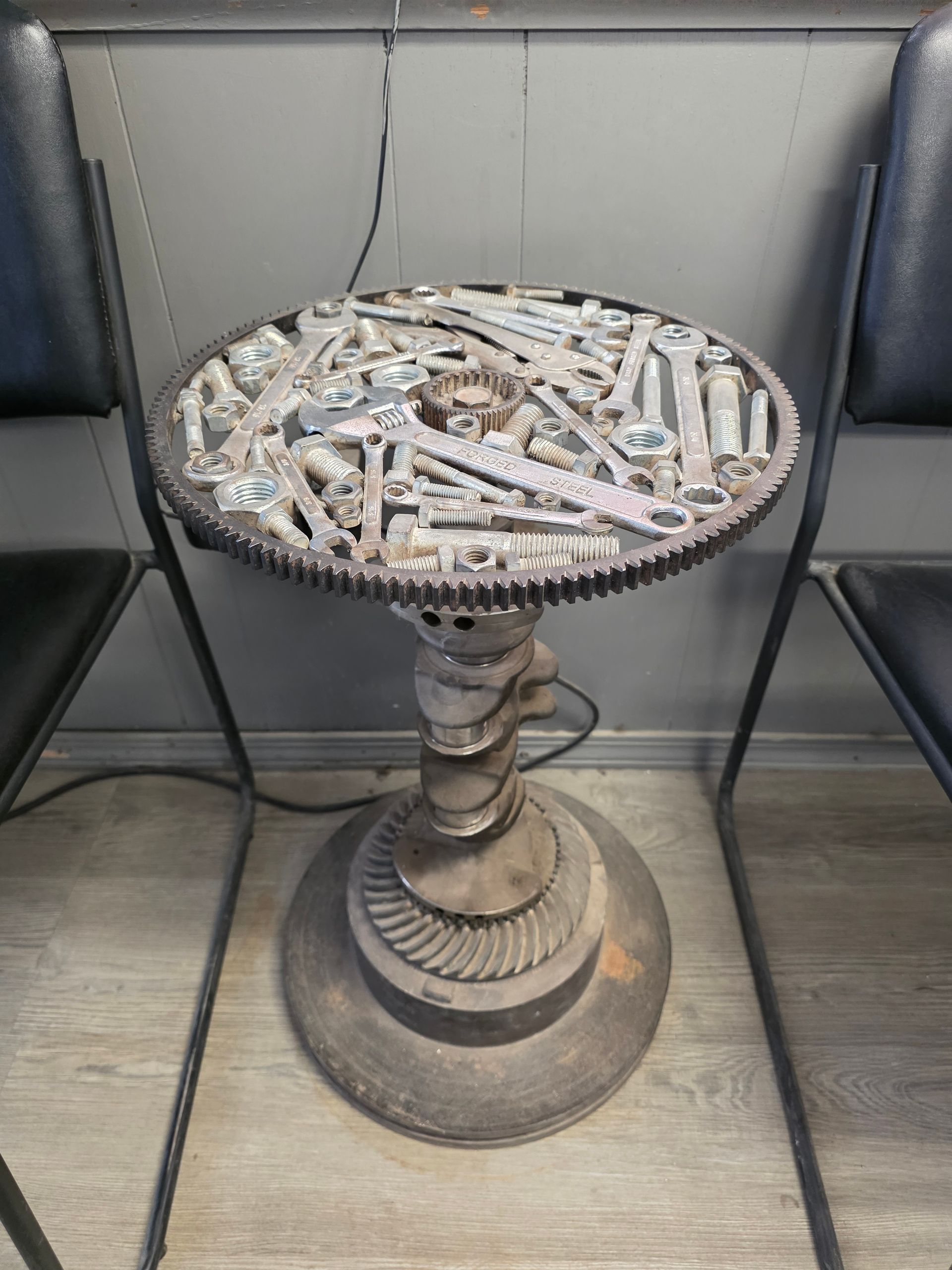 Table made from auto parts: gears, wrenches, and a circular top. Gray metal and dark chairs flank the table | Kaiser Automotive Tire & Alignment
