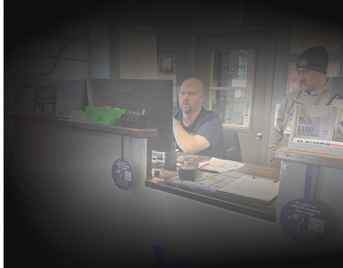 Woman smiling, sitting at a desk with computer monitors in an office. Wall signs visible.