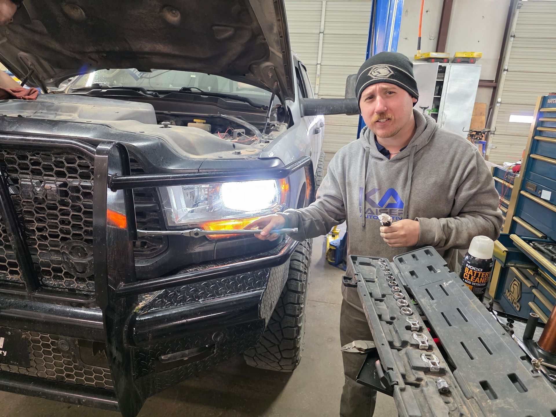 Man pointing at the headlight of a truck with the hood open in a garage, holding tools | Kaiser Automotive Tire & Alignment