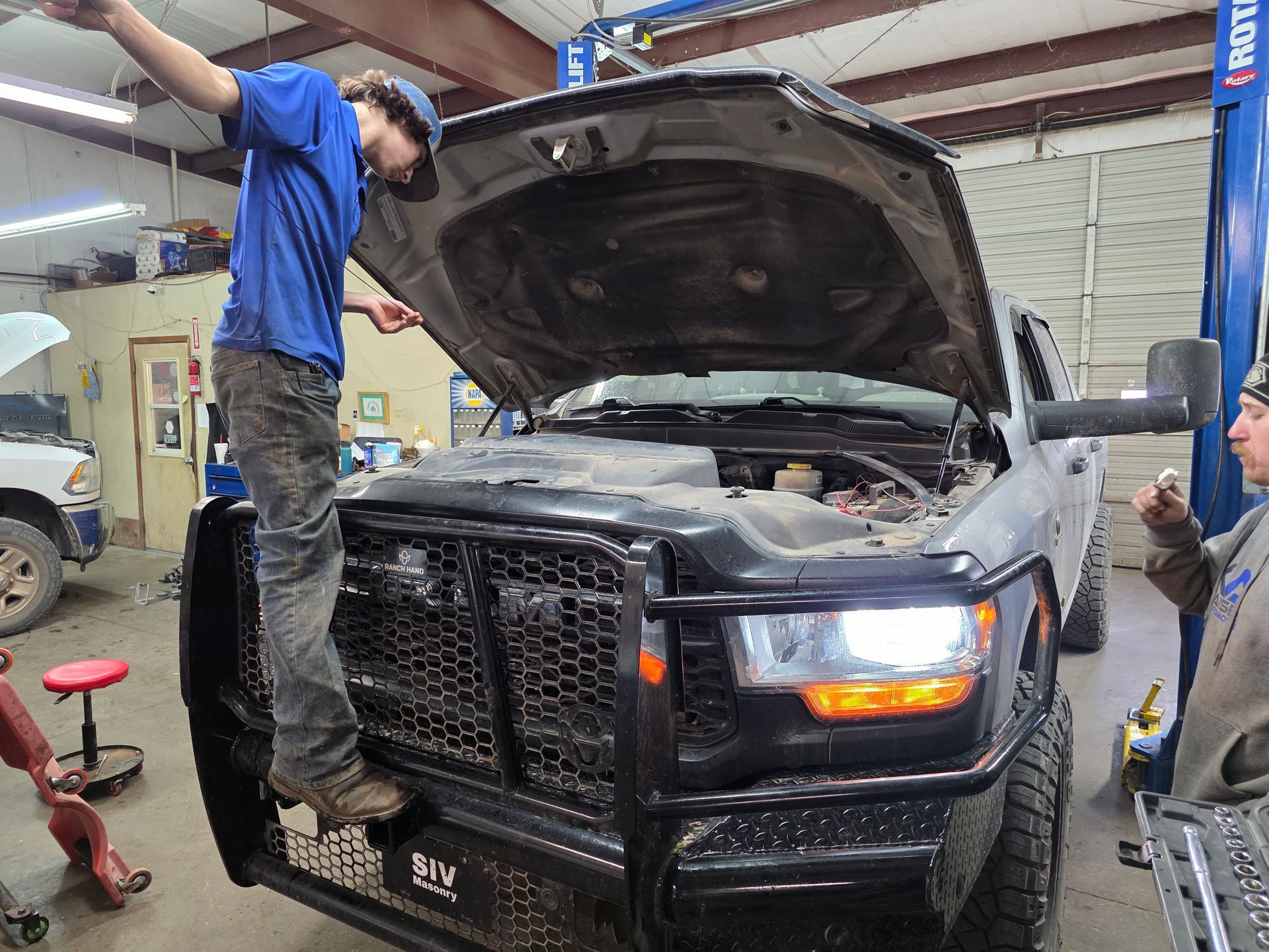 Mechanic on truck bumper, hood open. Another mechanic looks on in a garage setting | Kaiser Automotive Tire & Alignment