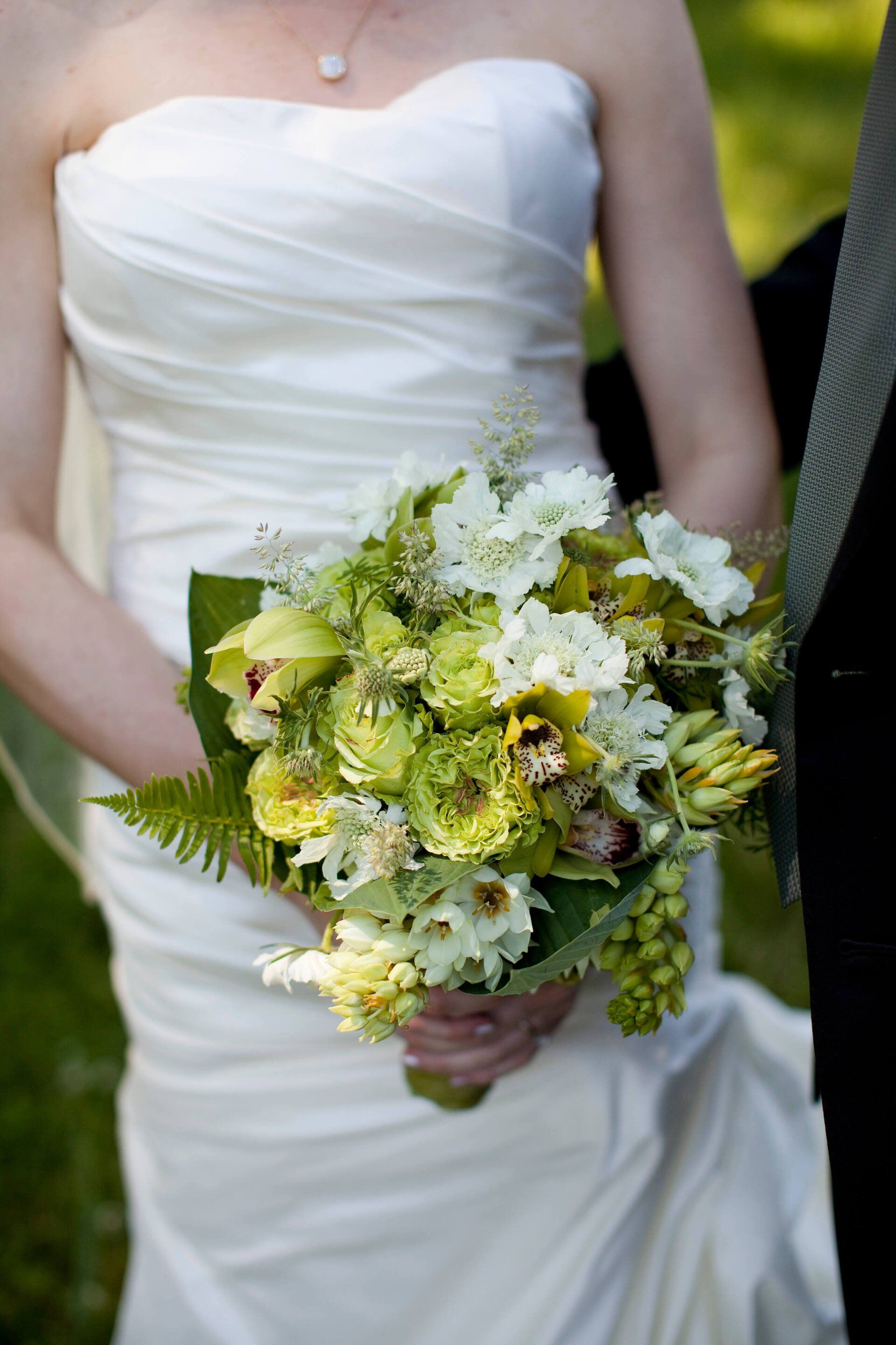 A bride in a white dress is holding a bouquet of green and white flowers - flowers designed by Flowers by Leo.