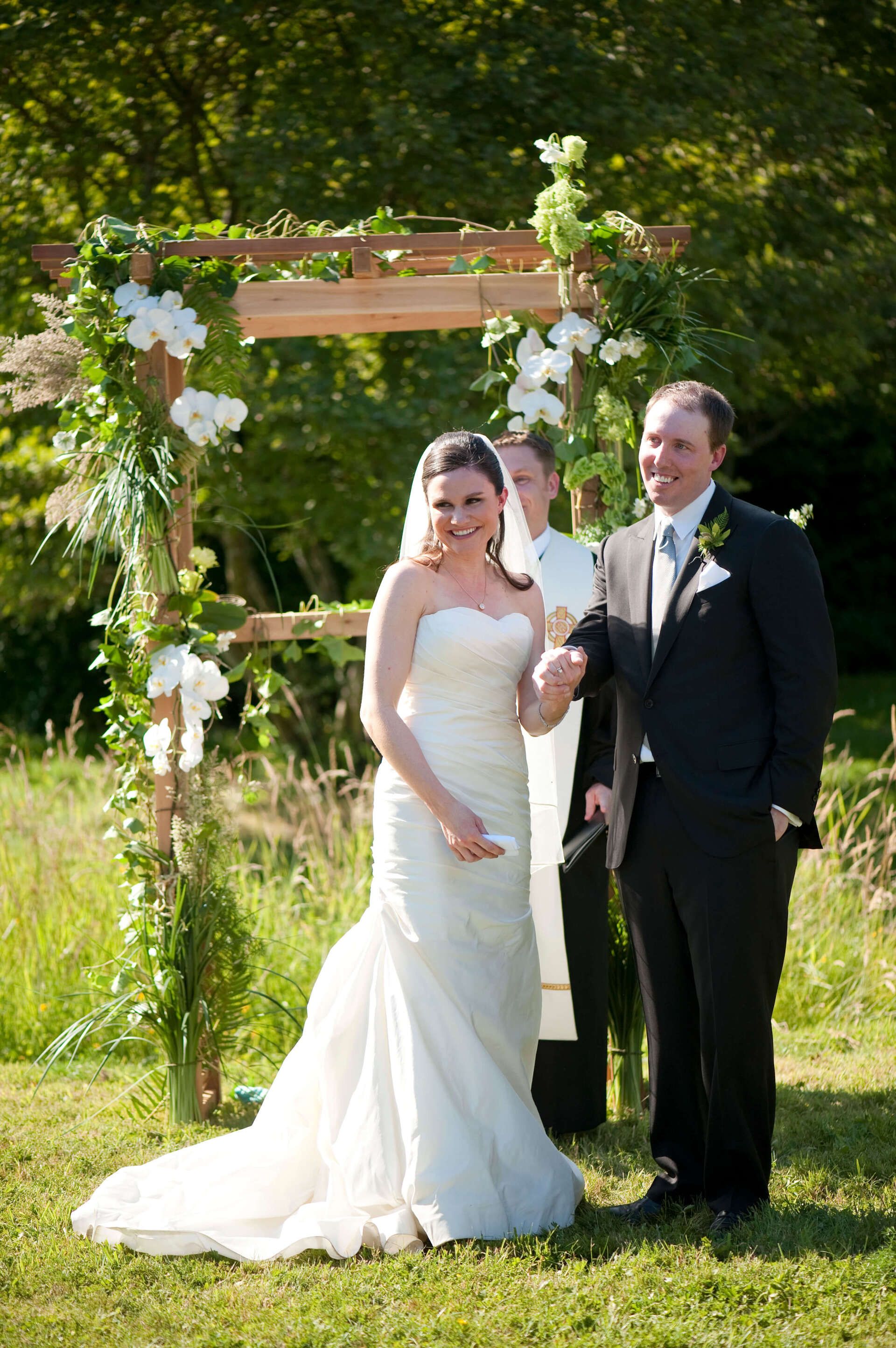 A bride and groom are posing for a picture in front of a wooden arch - flowers designed by Flowers by Leo.