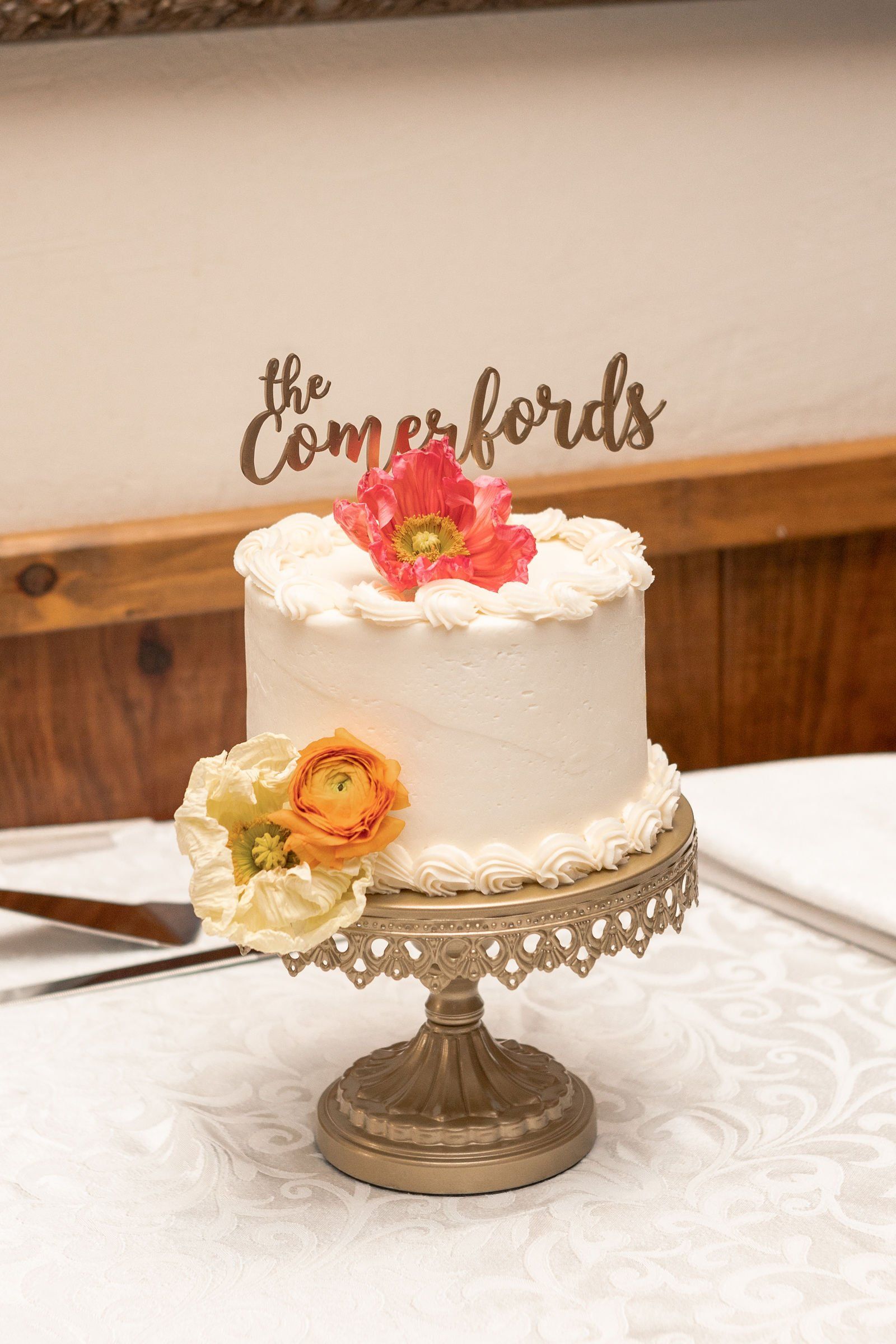 A small white cake with flowers on top is on a cake stand on a table - flowers designed by Flowers by Leo.
