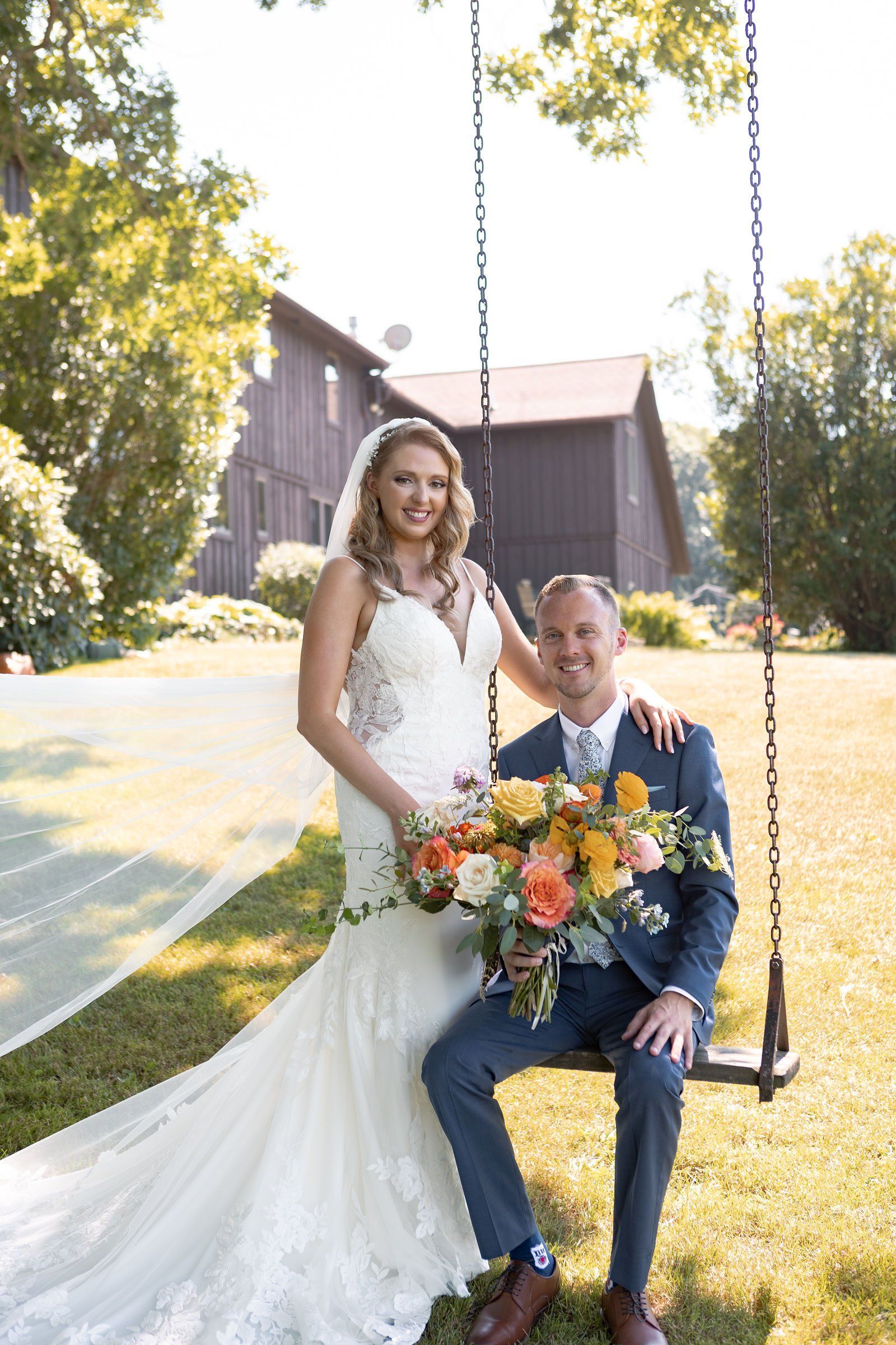 A bride and groom are posing for a picture while sitting on a swing - flowers designed by Flowers by Leo.