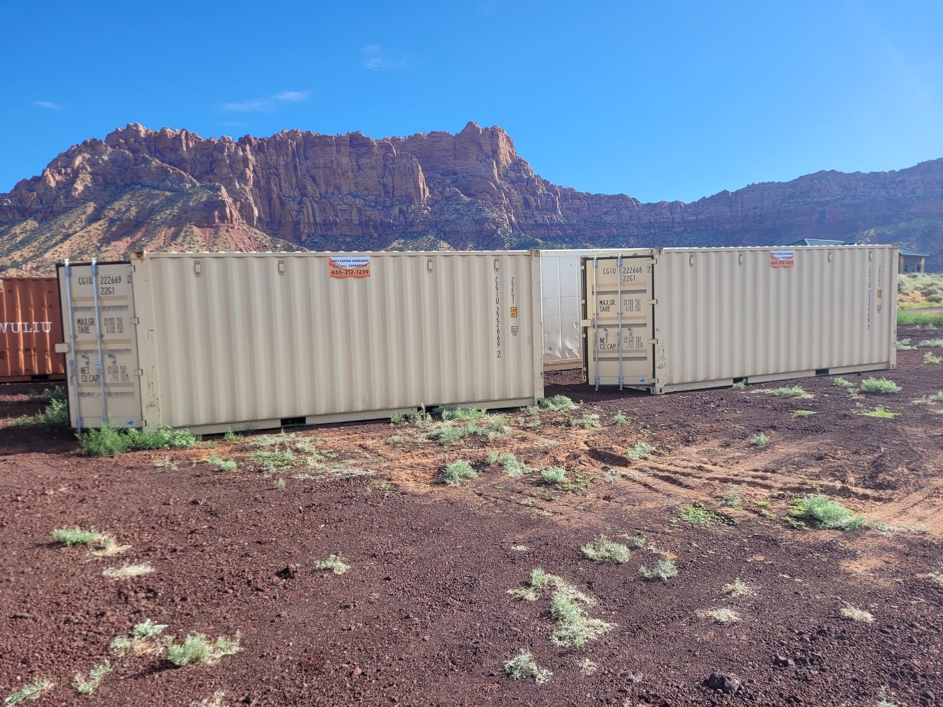 Two shipping containers are sitting in a dirt field with mountains in the background.