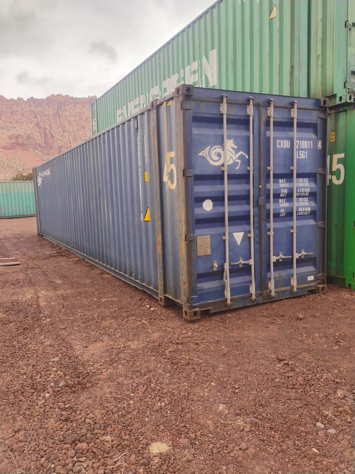 A blue shipping container is sitting in a dirt lot next to a green container.