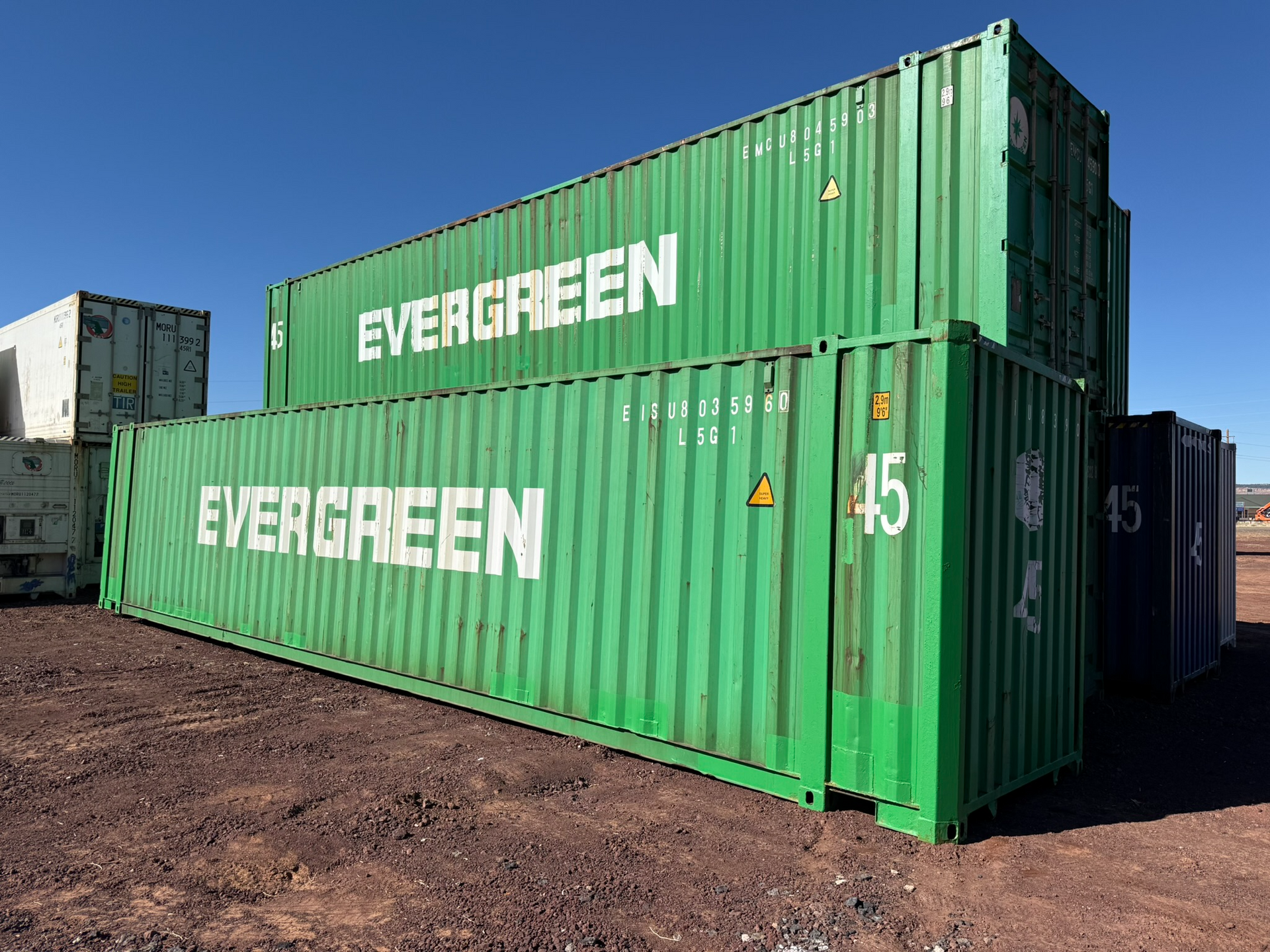Two green evergreen shipping containers are stacked on top of each other in a dirt field.