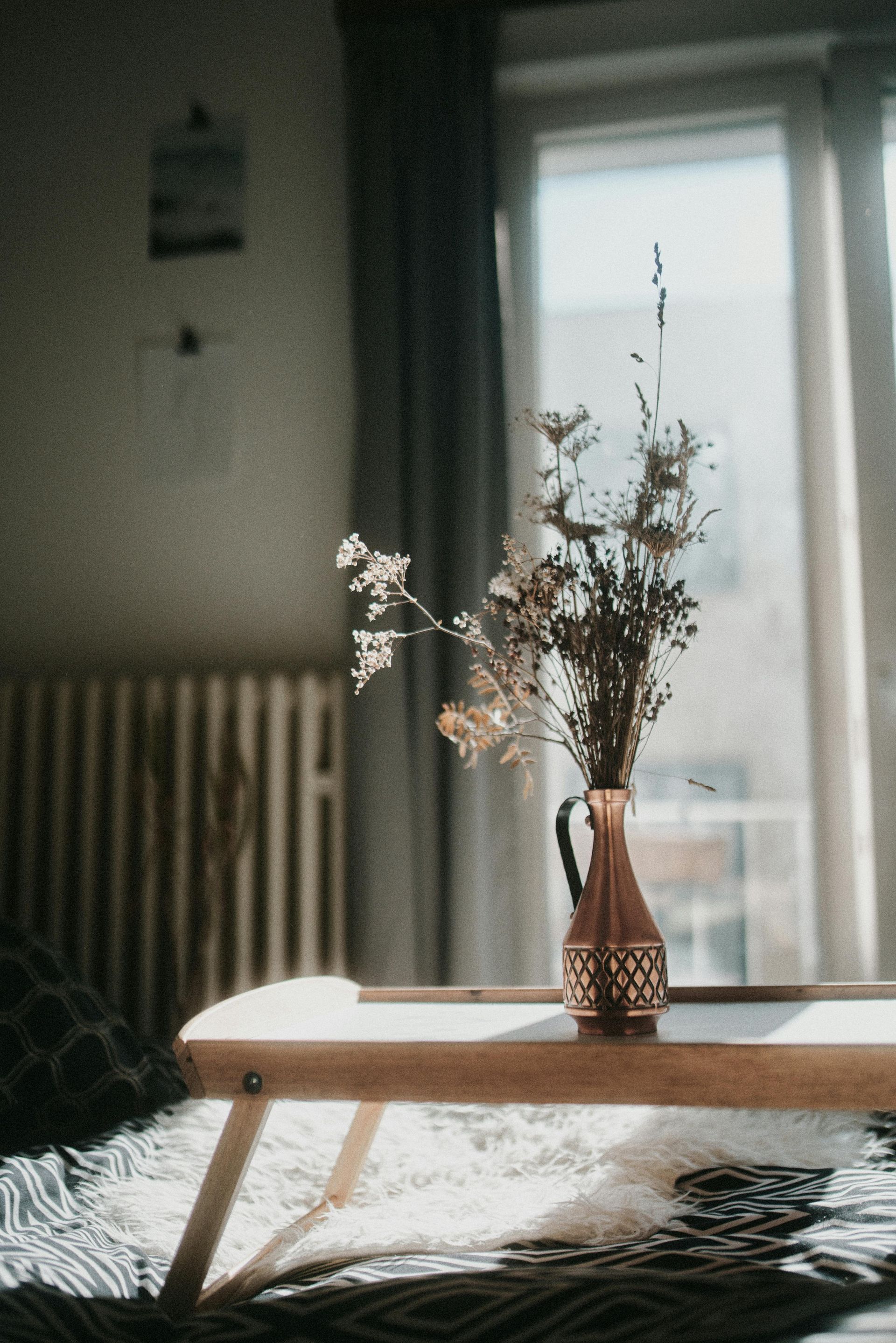 Dried flowers in a decorative vase on a wooden tray in front of a sunny window.