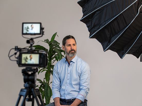 Man seated in a studio beside a camera and lighting umbrella, wearing a light blue shirt.