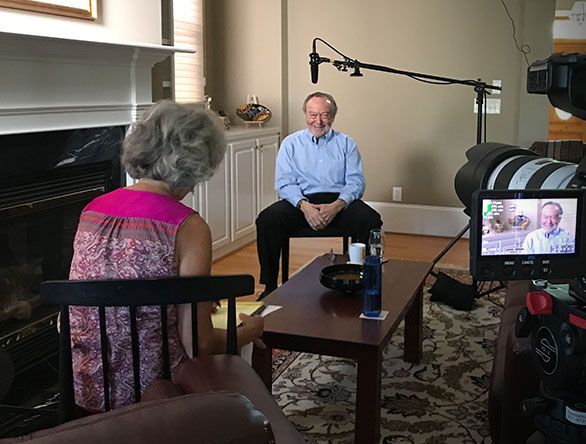 Two people sit in a living room being filmed for an interview, with a camera and boom mic visible.