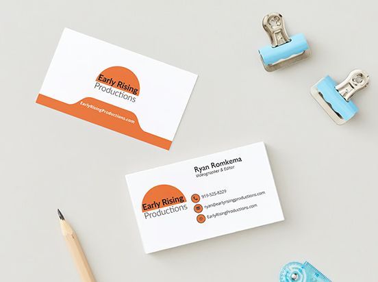 Business card mockup with orange-and-white branding on a light desk, with pencils and blue binder clips.