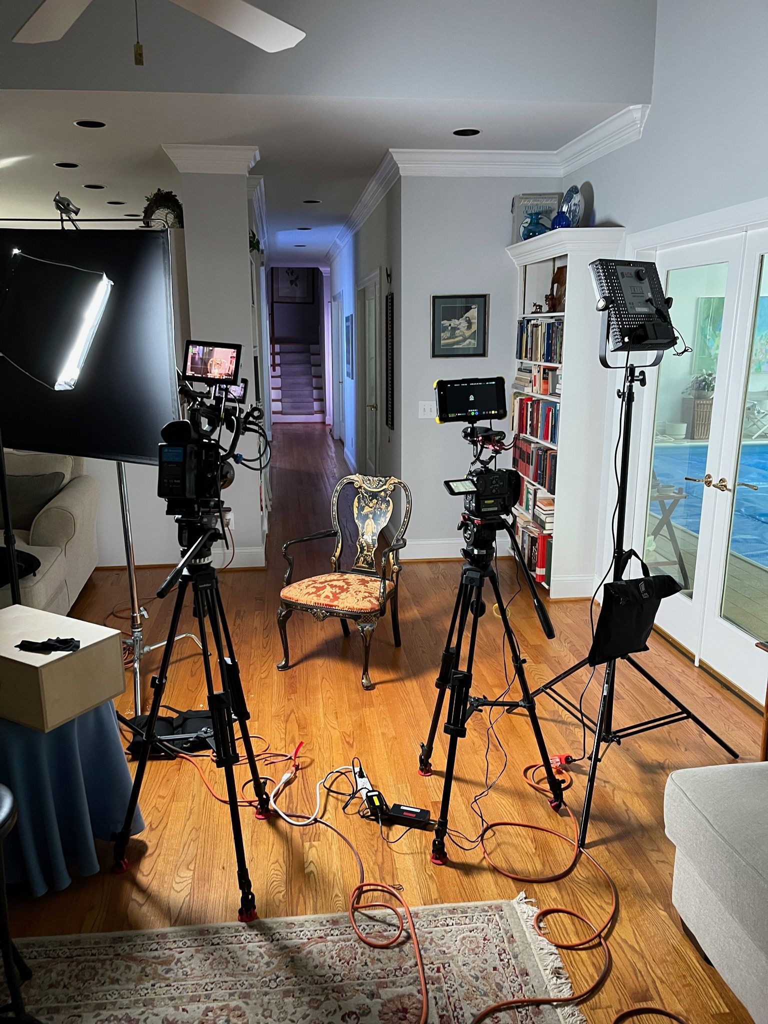 Home studio with multiple tripods, lights, and camera gear on a hardwood floor, viewed toward a hallway.