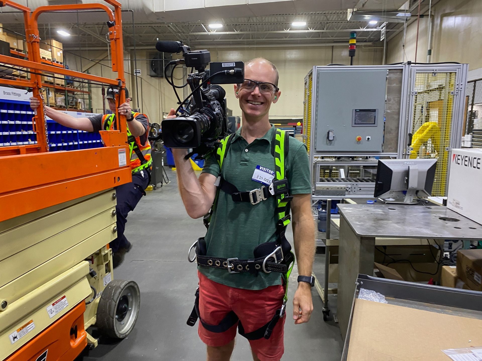 Smiling person holding a camera in an industrial workshop beside large machinery