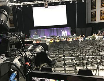 Conference hall with rows of chairs, stage with purple curtains, and a large camera in the foreground