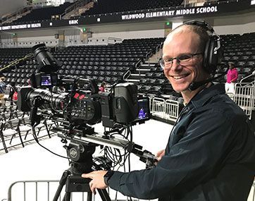 Camera operator with headset behind a broadcast camera in an empty gymnasium, smiling at the venue.