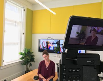 Person seated in bright studio, with a camera monitor in the foreground and a TV screen behind.