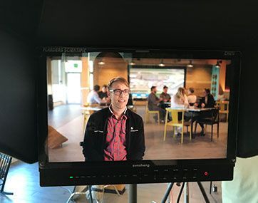 Man on a monitor in a café, with people seated at tables in the background.