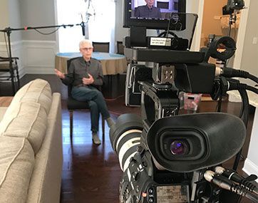 Camera in a living room filming a man seated in a chair near a sofa and TV screen.