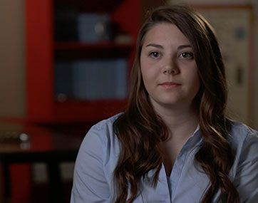 Woman with long brown hair in a light blue shirt, seated indoors against a blurred red-and-dark background