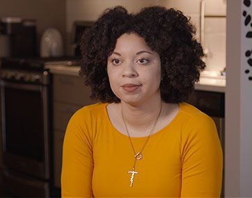 Woman with curly hair in a yellow top and cross necklace, sitting in a kitchen and looking at the camera.