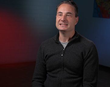 Man seated in a dark sweater, smiling against a red-and-gray studio background