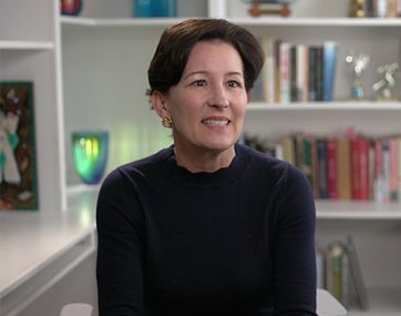 Woman in a black sweater seated in a bright office, smiling with shelves and books in the background