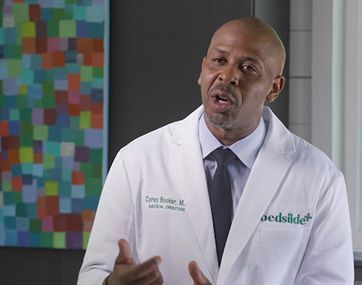 Doctor in white coat speaking in an office, with a colorful abstract painting in the background.