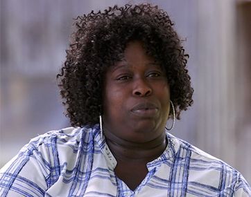 Person in a blue plaid shirt with curly hair, looking slightly off-camera against a blurred outdoor background.