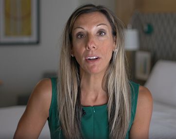 Woman in a green sleeveless top speaking indoors, with a blurred home background.