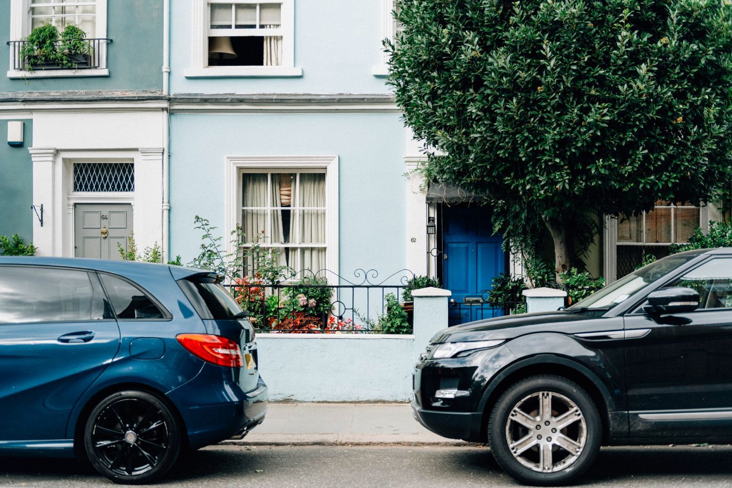 A blue car and a black car parked in front of a light blue townhouse with a large tree.