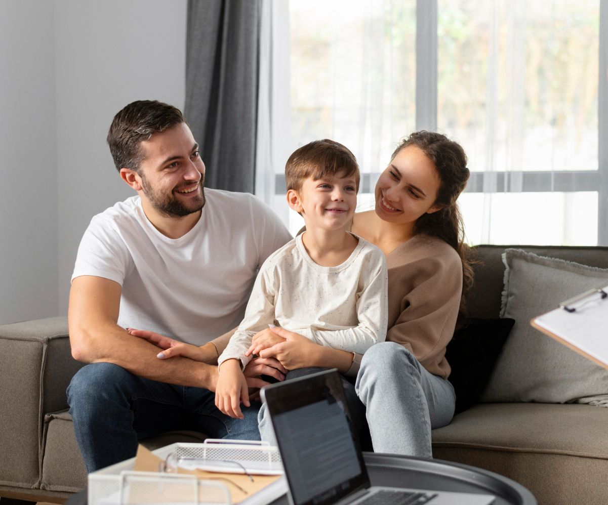 A family sits together on a couch, smiling and looking toward an off-camera person, with a laptop in the foreground.