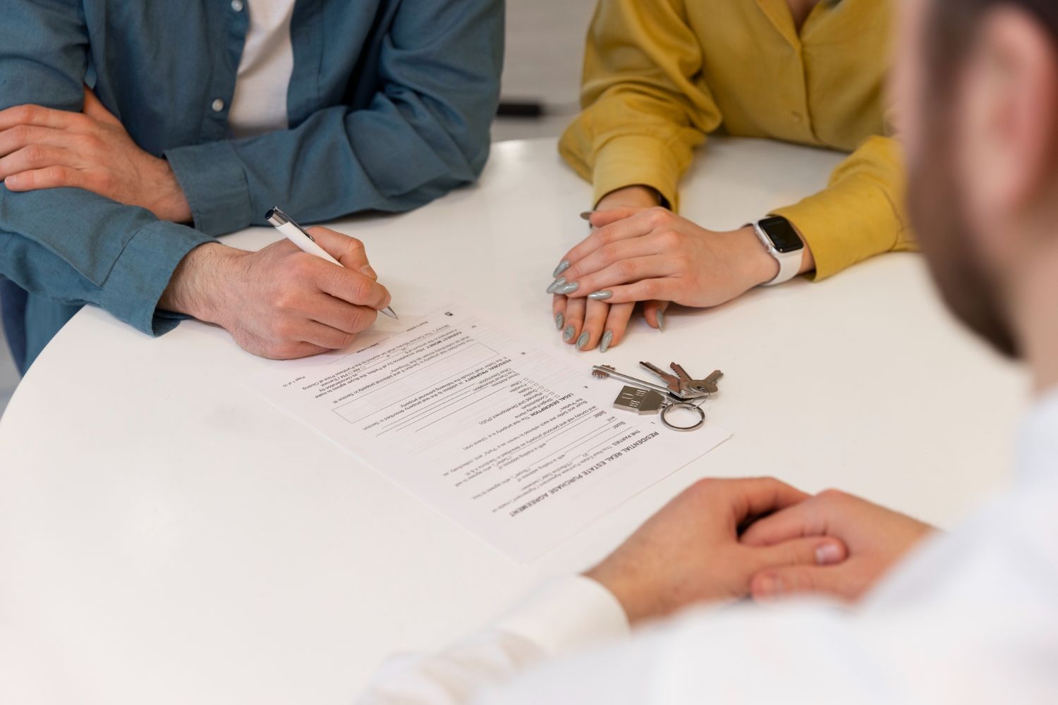 Two people sit at a table signing a document while a set of keys rests nearby, likely representing a real estate deal.