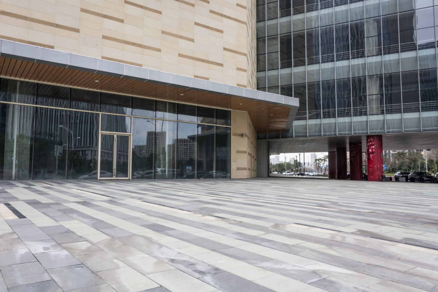 A modern building entrance with a glass facade, tan stone walls, and a grey paved plaza.