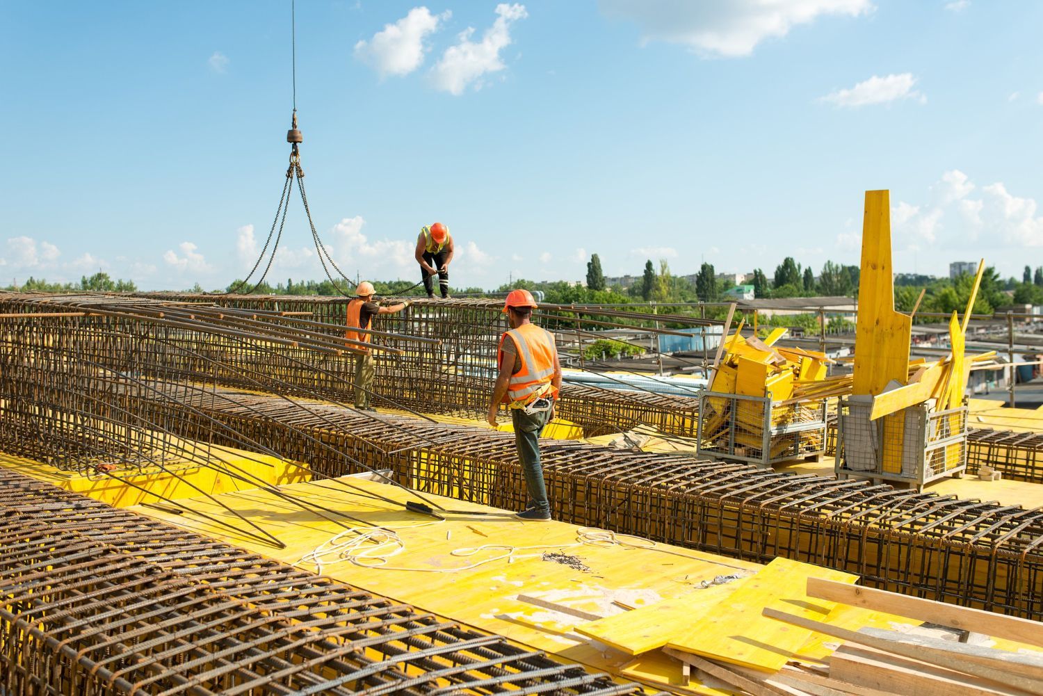 Construction workers assemble metal rebar frames on a bright yellow platform at an outdoor building site.