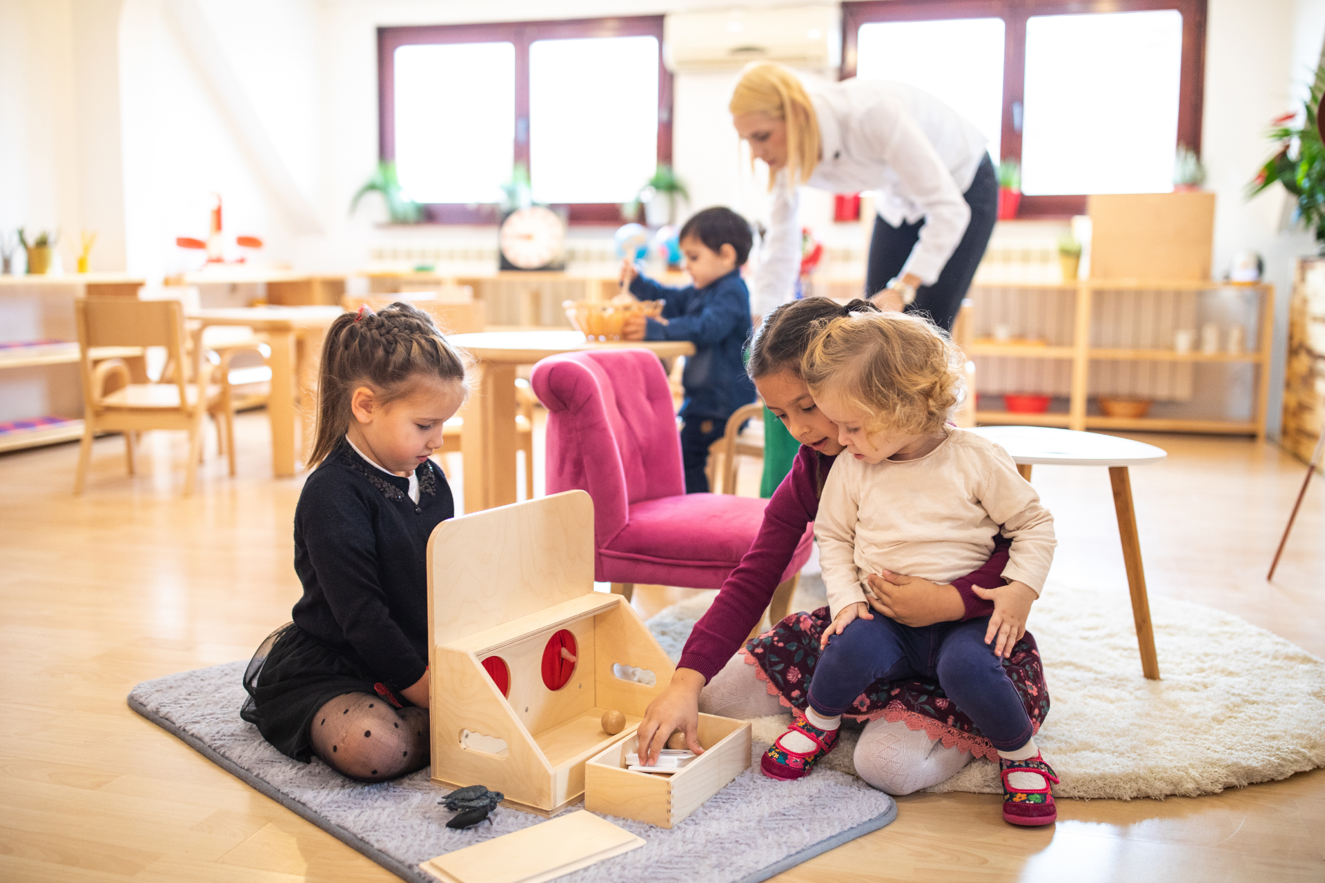 Children playing with wooden Montessori toys on a rug in a classroom, with a teacher supervising in the background.