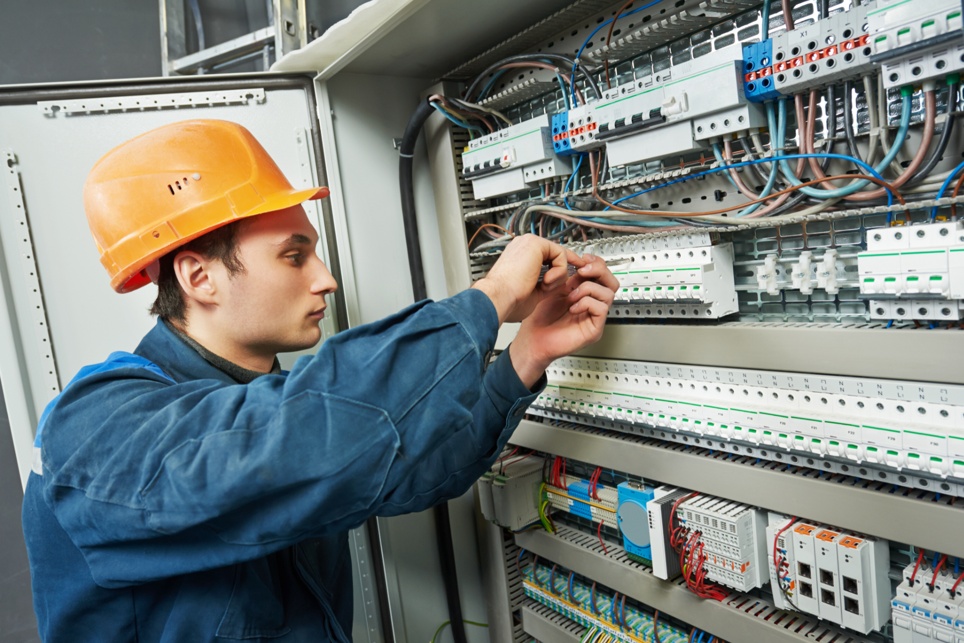 An electrician in a blue uniform and hard hat works on wiring inside a large industrial electrical control panel.