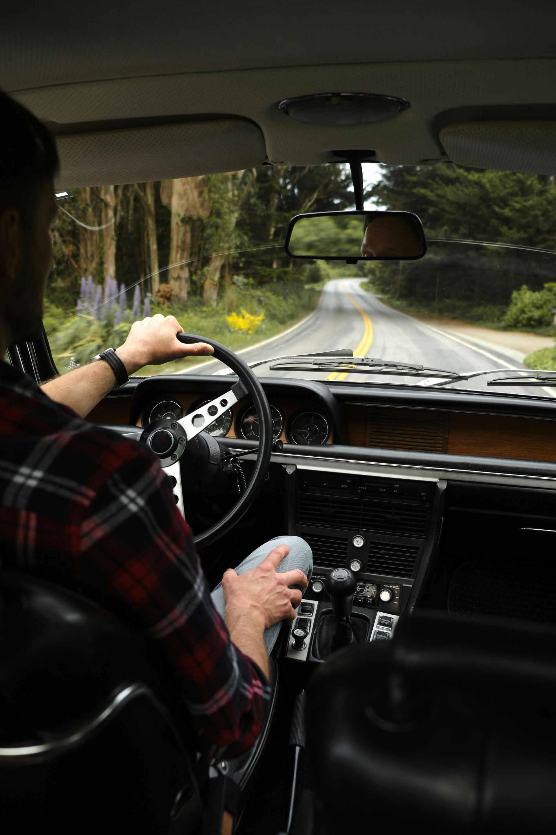 A person wearing a plaid shirt drives a vintage car along a scenic, tree-lined road.