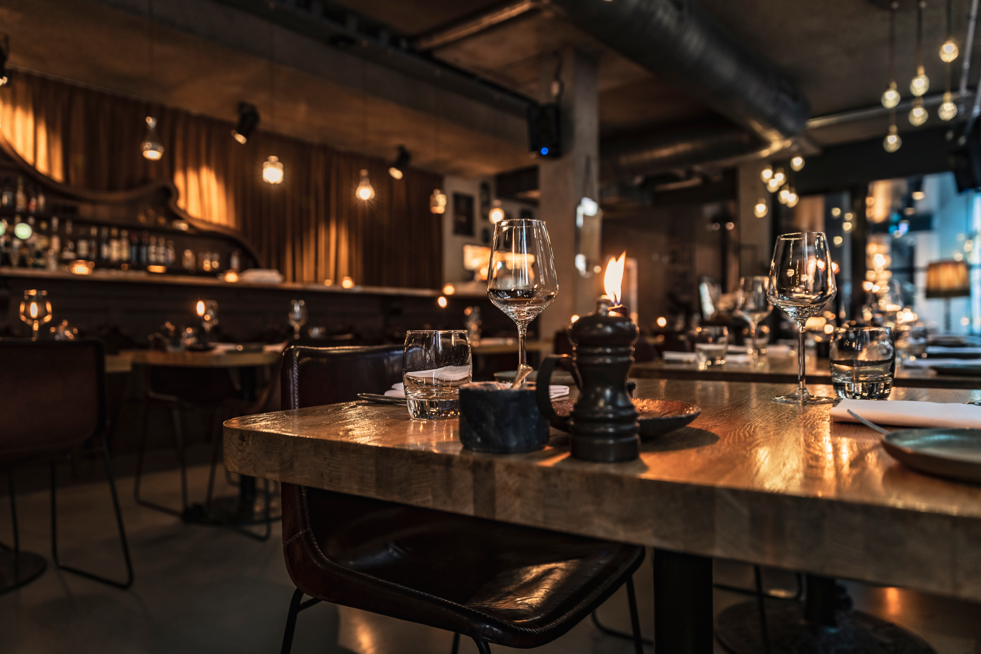 A warmly lit, dimly lit upscale restaurant interior focusing on a set wooden table with glassware and a pepper grinder.