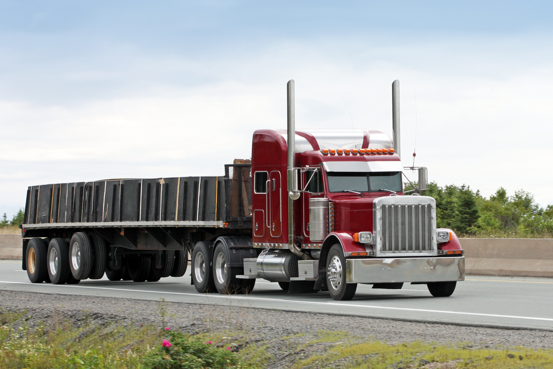 A red semi-truck with a flatbed trailer loaded with black metal sheets driving on a highway.