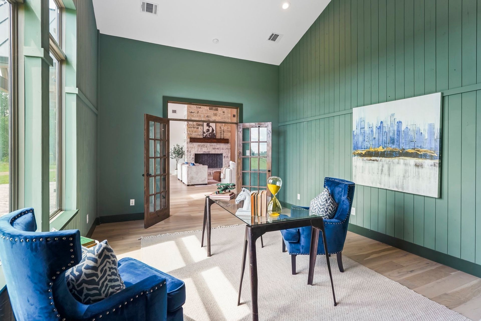 Green-walled home office with blue chairs, glass desk, and art.