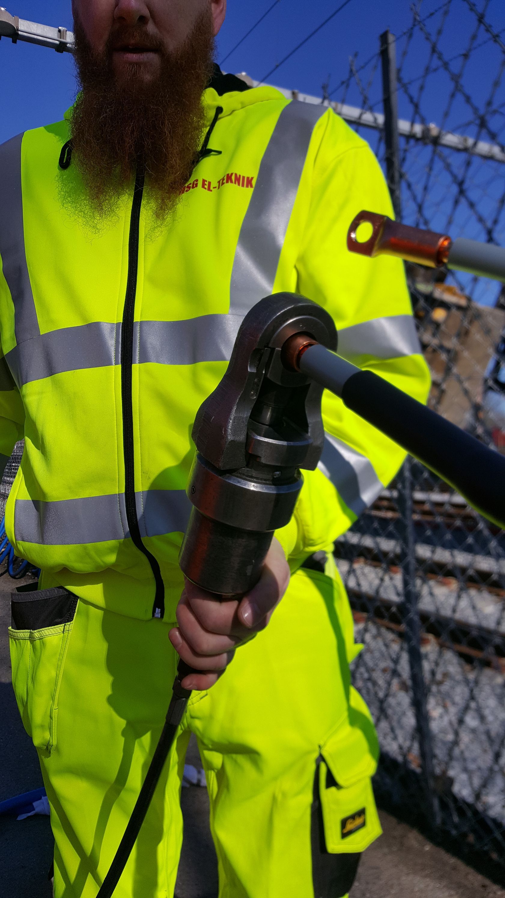 En mand med skæg har en gul sikkerhedsvest på og holder et værktøj.
