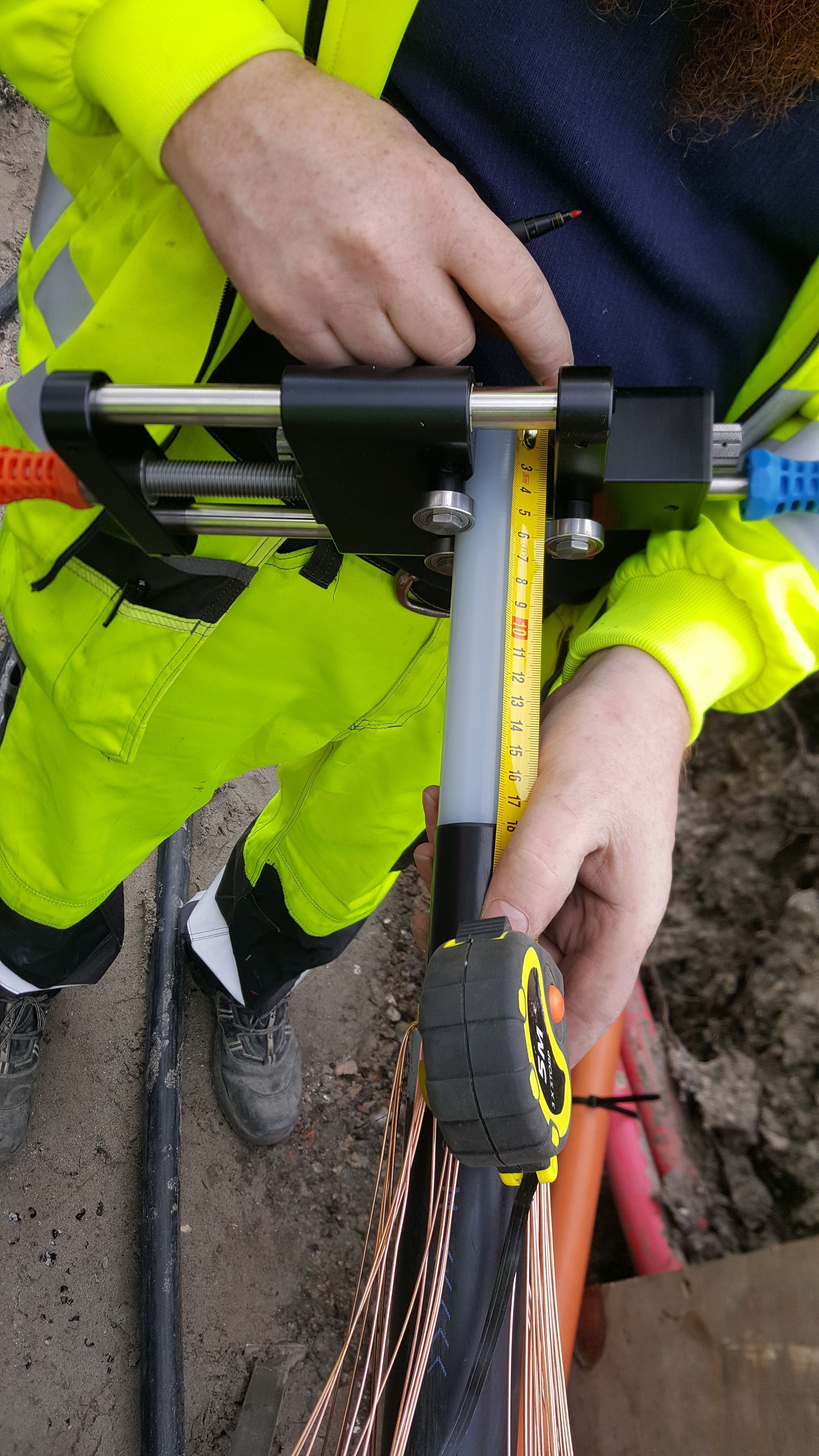 En mand i en gul jakke måler et rør med et målebånd.