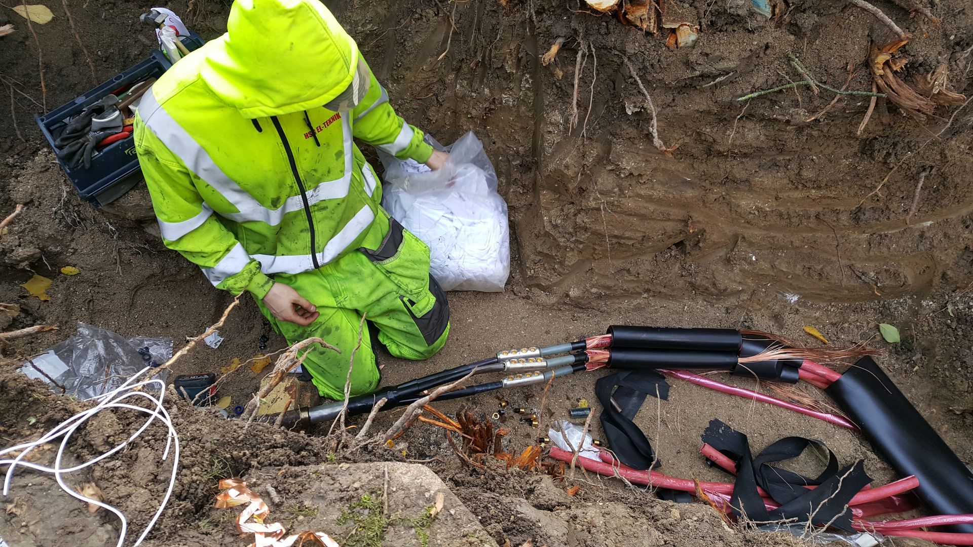 En mand i en gul jakke og grønne bukser knæler i jorden.