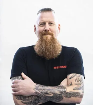 En mand med skæg og tatoveringer står med armene over kors.