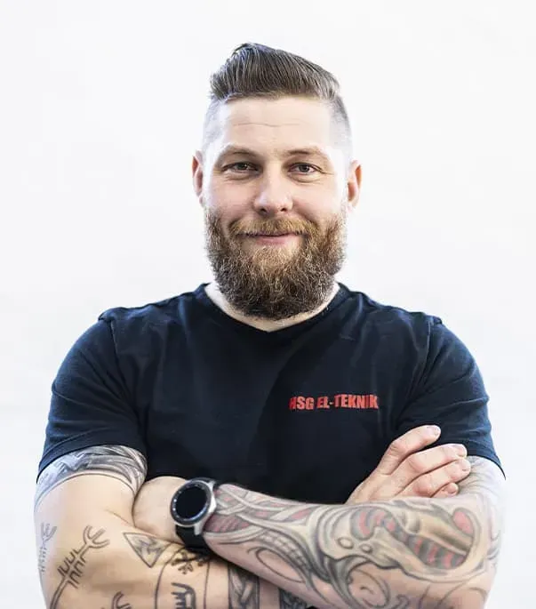 En mand med skæg og tatoveringer står med armene over kors.