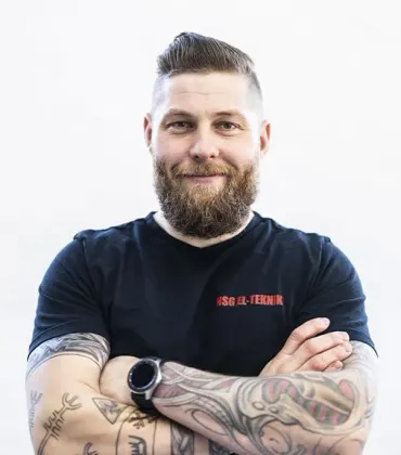 En mand med skæg og tatoveringer står med armene over kors.