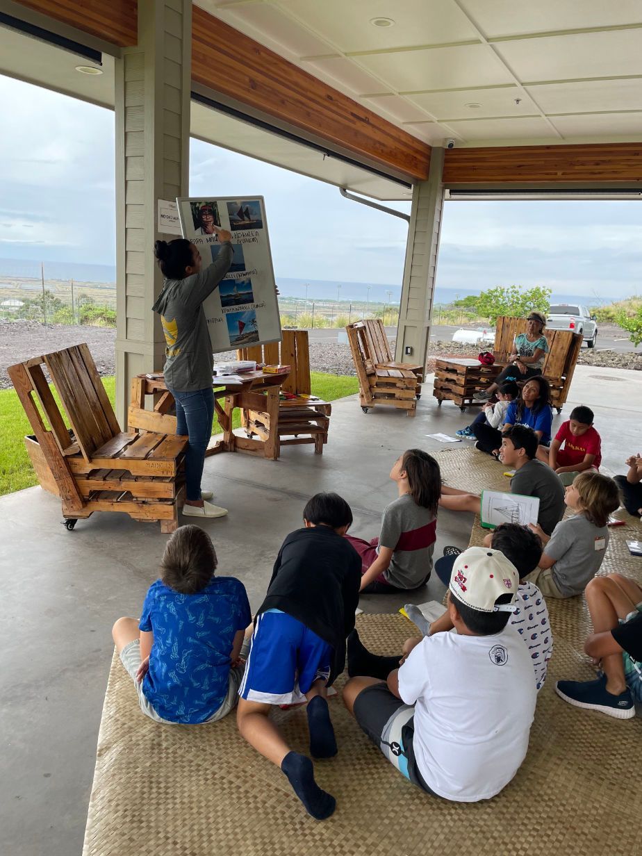 A group of children are sitting on the floor under a covered porch.